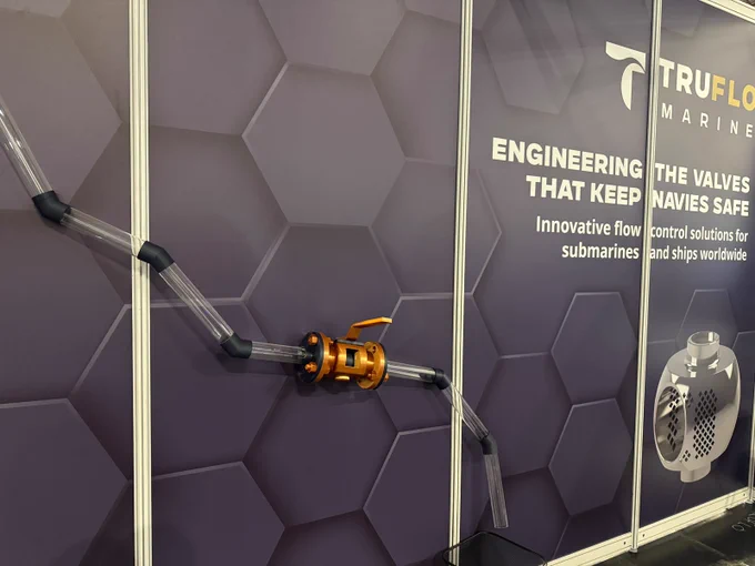 A wall display features clear tubing connected to an orange valve, showcasing TruFlo Marine. The backdrop includes text: "Engineering the valves that keep navies safe" and highlights offerings for submarines and ships worldwide. A hexagonal pattern enhances the design.