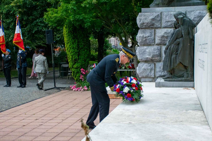 Vue de profil du préfet en uniforme déposant une gerbe de fleurs tricolore (bleu, blanc, rouge) au pied d'un monument aux morts en pierre. En arrière-plan, sur la gauche, des porte-drapeaux et d'autres personnes en uniforme ou en tenue civile assistent respectueusement à la cérémonie dans un parc arboré.