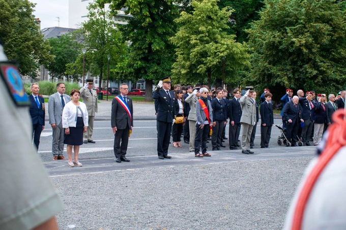 Une rangée d'officiels, de militaires en uniforme et d'élus se tiennent au garde-à-vous et saluent lors d'une cérémonie commémorative en plein air. En arrière-plan, un groupe de jeunes et d'anciens combattants participent à la cérémonie devant des arbres.