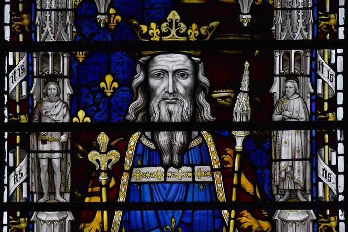 Edward III depicted in a stained glass window. The King wears and gold crown and red and blue robes, and holds a sceptre in each hand. He has long hair and a long beard.