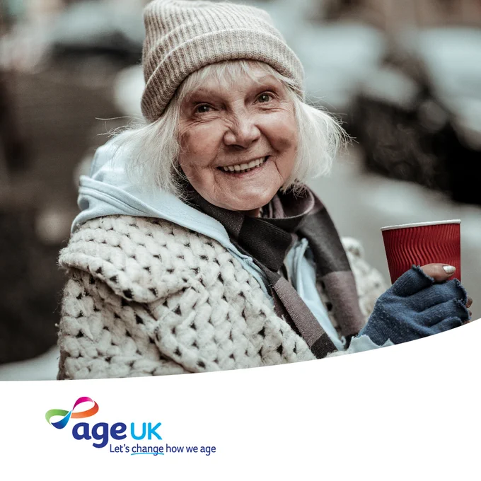 An older adult wearing a knitted hat and a scarf smiles while holding a red cup. The person is dressed warmly with gloves and a thick coat. The Age UK logo and the tagline "Let's change how we age" are displayed at the bottom of the image.