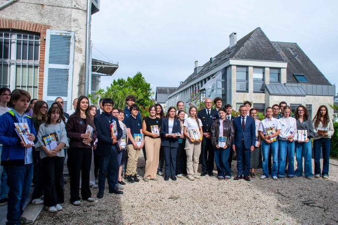Photo de groupe officielle en extérieur réunissant les lauréats tenant fièrement leurs livres reçus en récompense. Au centre du groupe se tiennent le préfet des Hautes-Pyrénées et le secrétaire général de la DSDEN. Ils posent tous ensemble dans la cour en gravier d'un bâtiment officiel.