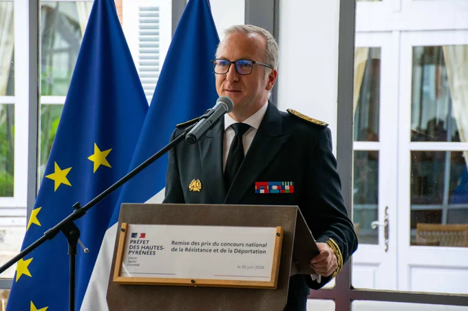 Le préfet des Hautes-Pyrénées s'exprime derrière un pupitre équipé d'un microphone. Sur le pupitre, une affiche officielle indique : « Remise des prix du concours national de la Résistance et de la Déportation - le 05 juin 2026 ». Les drapeaux européen et français sont visibles en arrière-plan.