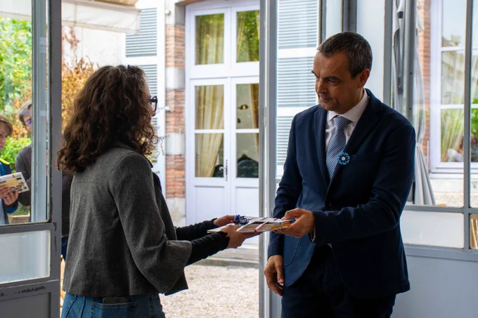 Le secrétaire général de la DSDEN remet un prix composé de livres à une jeune fille. La scène se déroule près d'une grande porte vitrée ouverte donnant sur l'extérieur.