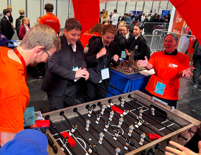 A group of people enthusiastically play foosball at a UK Power Networks booth. They are surrounded by onlookers and exhibitors in a busy event setting.