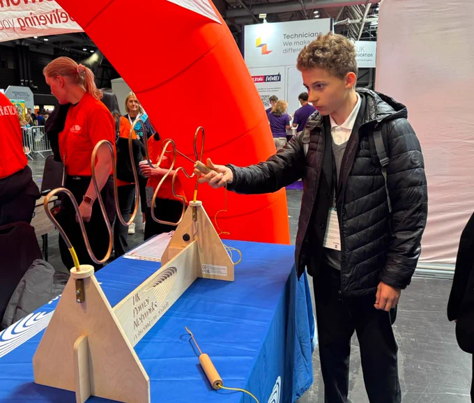 A person is interacting with a wire loop game at the UK Power Networks booth. The setup includes a curved metal wire and a handheld loop. The booth has a blue tablecloth and a large red inflatable structure as part of the display. The background features banners and another person.