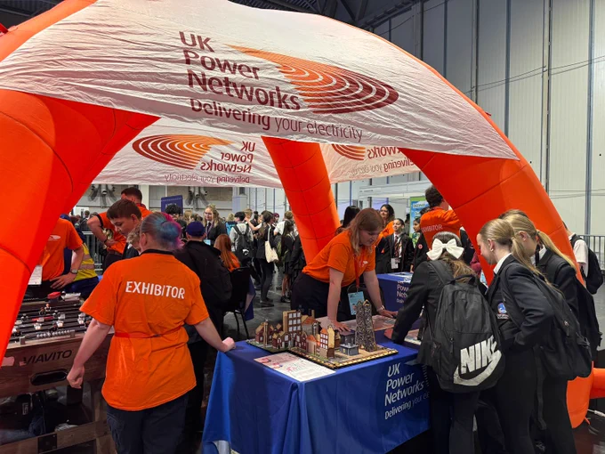 A UK Power Networks booth with an orange canopy displaying their logo and slogan. People are interacting with exhibits, including a model display on a table. Some individuals wear orange shirts labeled "Exhibitor." Attendees are engaged around the booth.