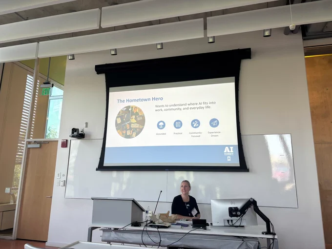 Jessica Stansbury sits at the front of a modern classroom during an AI summit presentation at the University of Baltimore. Behind her, a large projection screen displays a slide titled “The Hometown Hero."