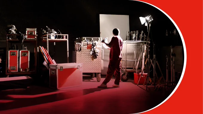 Technician adjusting lighting equipment on a dark film set surrounded by gear cases and stands.