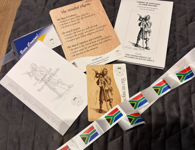 Camino pilgrimage items laid out, including a pilgrim passport, guidebook, blessing card, badge, and South African flag ribbons.