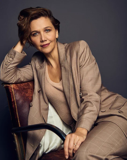 Maggie Gyllenhaal in a taupe suit sitting on a chair smiling at the camera
