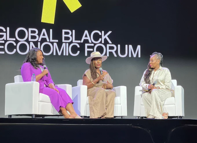 Three Black woman sit on chairs in front of a screen that reads "Global Black Economic Forum"