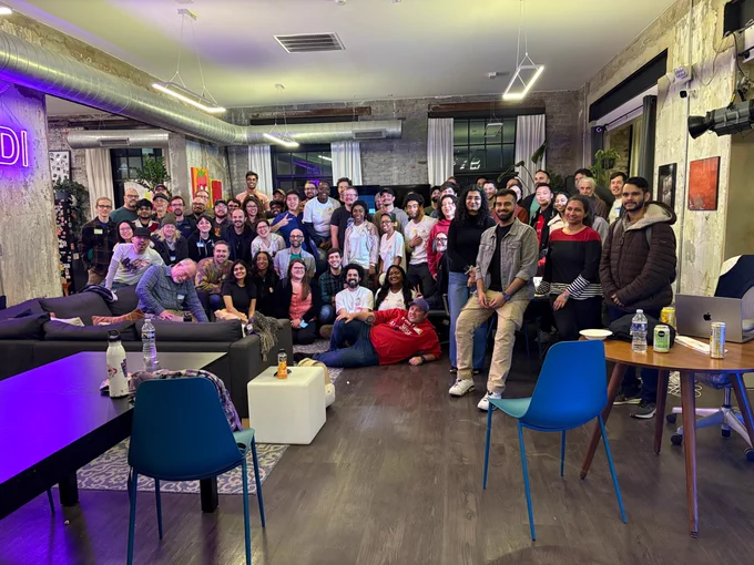 A large group of people poses together indoors in a modern, well-lit room with sofas, tables, and bright artwork on the walls.