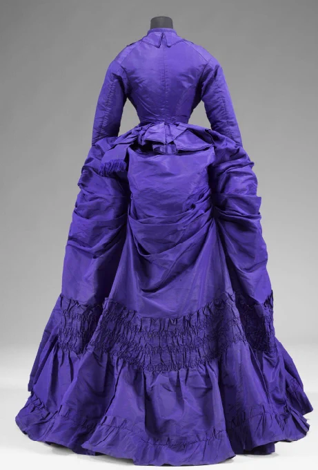 Back view of the early 1870s purple silk bustle dress which shows the full complexity of the bustled skirt with its many drapes and ruches. The bodice is long sleeved and fitted