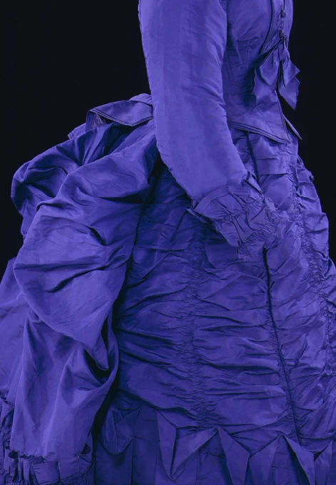 Partial view of the side of a bright purple bustle dress. It has long sleeve and plentiful ruching down the skirt with piped seams and a fringed bow