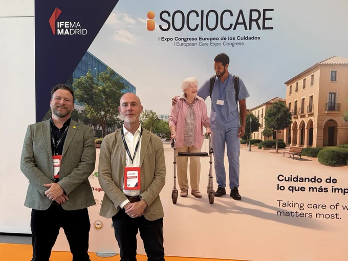 Two men in business attire stand in front of a SOCIOCARE conference backdrop at IFEMA Madrid, promoting elderly care.