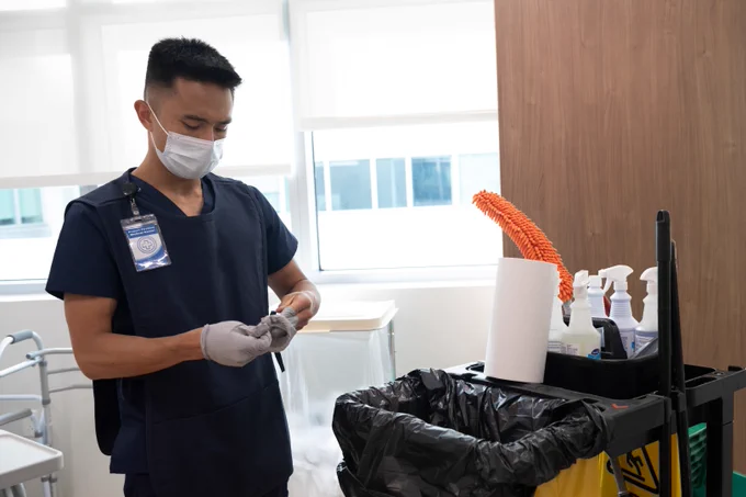 EVS worker wearing blue scrubs and mask is taking off gloves in front of a cleaning cart.