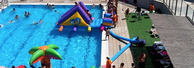 Piscina al aire libre con un inflable acuático tipo circuito de obstáculos y varias personas jugando y tomando el sol.