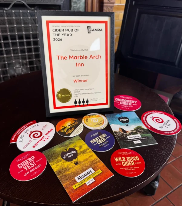 Central Manchester CAMRA branch certificate awarding Cider Pub of The Year to The Marble Arch Inn. Its frame sits on a polished, dark wood table alongside lots of brightly coloured cider clips from independent producers.