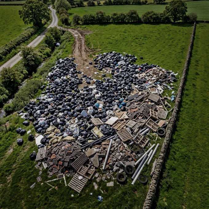 A rural landscape showing a large pile of dumped rubbish, including black bags, wooden pallets, tyres, and construction debris, on a green field bordered by stone walls. A road curves around the left side, with trees and hedges lining the area.
