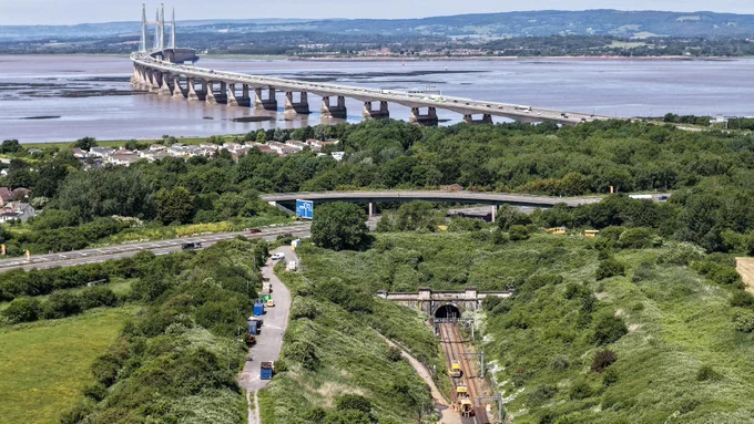 Scenic view of a bridge spanning a wide river, surrounded by lush green landscape. In the foreground, roads and railway tracks lead toward a tunnel.