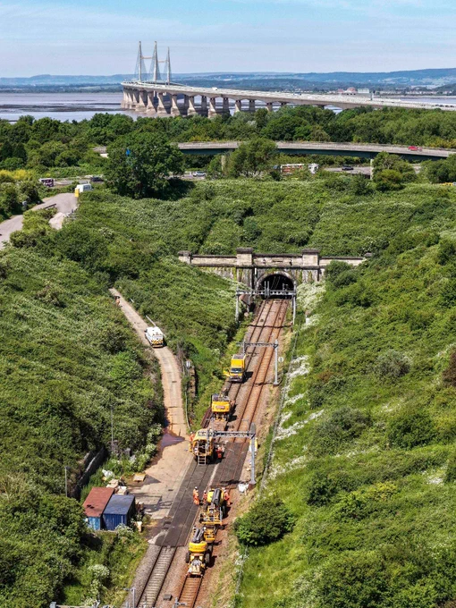 An aerial view showing railway maintenance vehicles working on tracks leading into a tunnel surrounded by greenery. In the background, the Severn Bridge spans across a body of water.