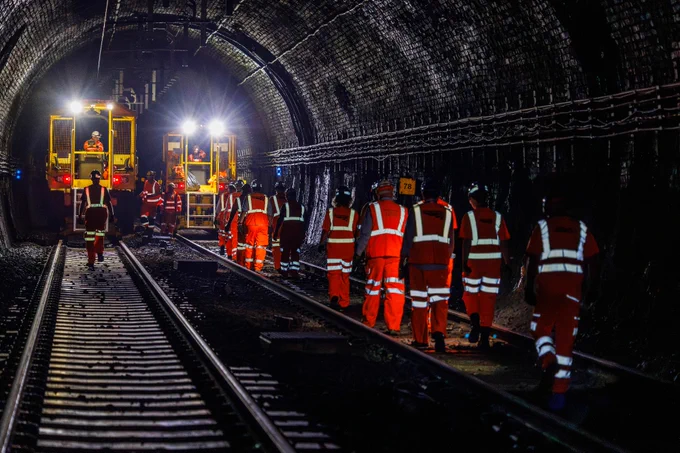 Workers in high-visibility clothing are walking through a dimly lit tunnel alongside train tracks. Bright lights illuminate equipment in the background.