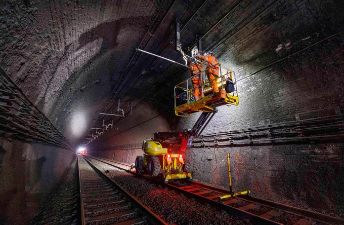 This image shows a worker on an elevated platform vehicle inside a dimly lit railway tunnel, performing maintenance on overhead cables. The tunnel's brick interior and railway tracks are visible, leading into the distance.