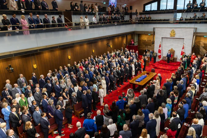 Wide view of a parliamentary chamber filled with standing attendees, with a red carpeted floor and a central table holding ceremonial items, as officials and uniformed guards stand near a raised dais flanked by Canadian flags.