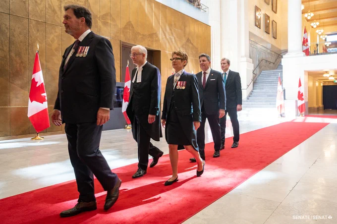 Five formally dressed individuals walk in single file along a red carpet inside a grand interior hall, with Canadian flags displayed along the sides and medals visible on several jackets; a staircase, warm-toned walls, and ceremonial setting  appear in the background.