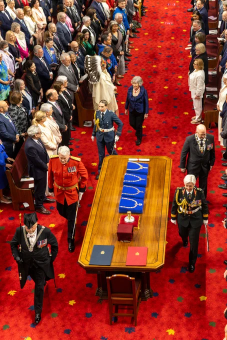 Overhead view of a formal parliamentary chamber with rows of seated and standing attendees lining both sides of a red carpeted aisle, as four uniformed officials and two formally attired individuals walk toward a central wooden table holding ceremonial items.