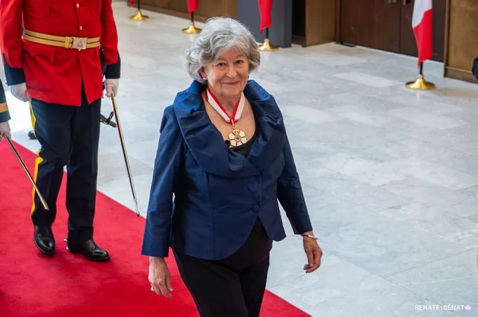 A woman wearing a navy blazer and a medal walks along a red carpet inside a formal setting, with another uniformed individual in a red ceremonial uniform following behind; Canadian flags and a light stone floor are visible in the background.