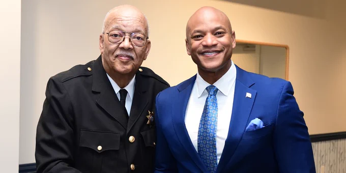 Two men shaking hands indoors, one dressed in a formal police uniform and the other in a blue suit with a patterned tie and pocket square.