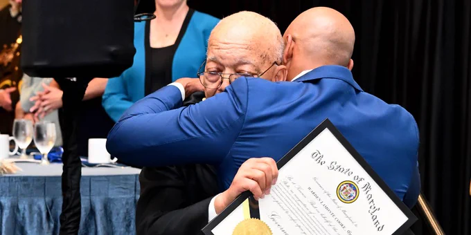Two men embrace warmly as one holds a framed Senate resolution during a formal event with a blue-draped table and attendees in the background.