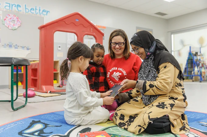 Alt text: An instructor wearing a red RRC Polytech shirt sits on a classroom rug with two young children and an adult caregiver wearing a patterned hijab. The group is smiling and looking at a book together in an early childhood education setting.