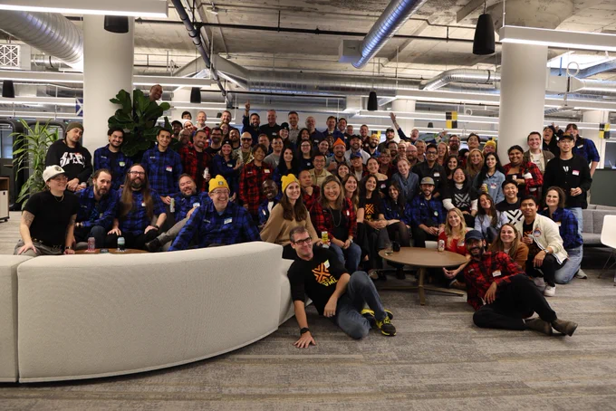 A large group of people is gathered in a modern office space with industrial design elements, such as exposed ducts and pillars. Many are wearing plaid shirts and some have yellow beanies. The group is smiling and appears to be casually posing together.