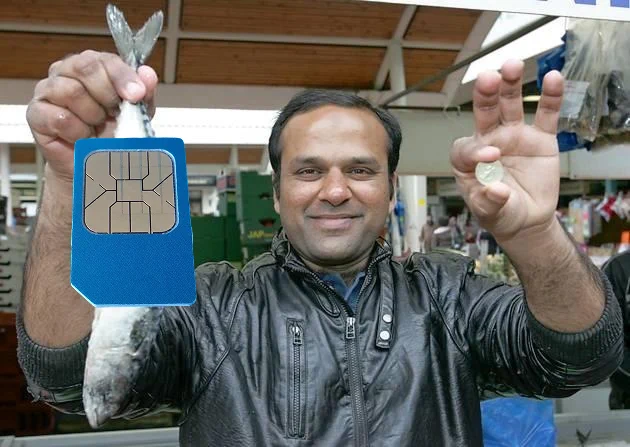 The internet sensation Muhammad Shahid Nazir, also known as the One Pound Fish Man, holding up a fish in his right hand and a one pound coin in his left hand. Except a blue Sim card has been superimposed on top of the fish.