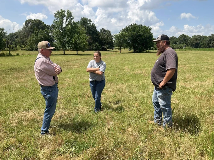 Three people standing in a grassy field under a partly cloudy sky, engaged in conversation with arms crossed.