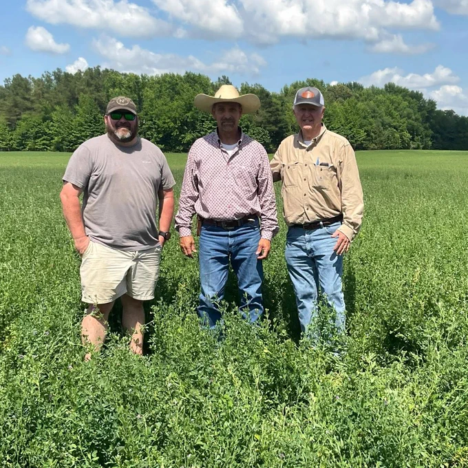 Three men standing in a lush green field under a partly cloudy sky, dressed casually with one wearing a cowboy hat.