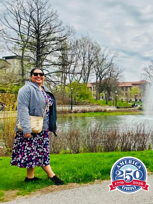 A person stands smiling near a pond with a water fountain. Trees and buildings are in the background. They are wearing sunglasses, a floral dress, and a gray jacket, holding a beige bag. A "America 250 Ohio 1776 2026" logo is visible in the bottom right corner.