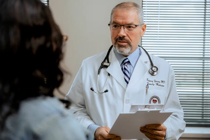 Doctor in white coat with stethoscope holding a clipboard, speaking to a patient.