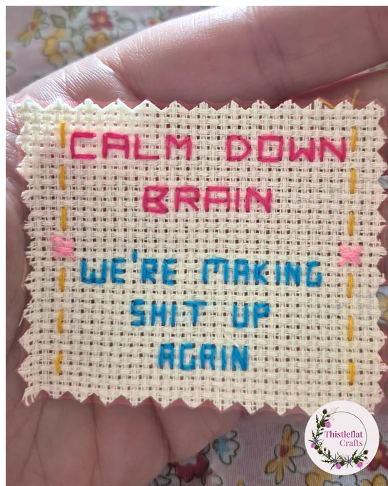 Small 2 x 2" piece of aida with the quote back stitched in bright pink and turquoise 'Calm down brain. We're making shit up again'
