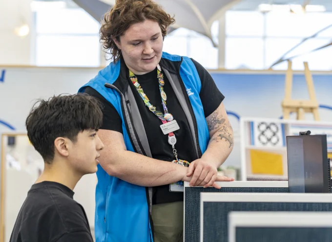A person wearing a blue vest assists another individual seated in front of a computer.