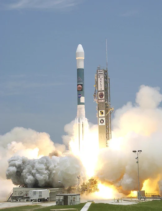 A rocket is launching into the sky, surrounded by clouds of smoke and bright flames. The rocket bears the logos of NASA and Delta II. The launch pad is visible with a clear sky in the background.