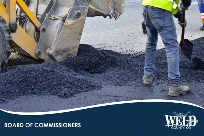 Construction worker spreading fresh asphalt on a road next to a backhoe loader during paving work.