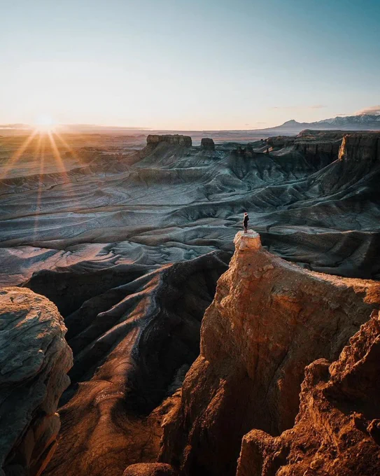 A lone person stands atop a narrow sandstone pinnacle overlooking Moonscape Overlook in Utah at sunrise. Gray bentonite badlands ripple across the landscape like an alien world, with deep canyons, towering cliffs, and distant mountains stretching to the horizon. Golden sunlight spills across the scene, emphasizing the immense scale of the rugged terrain.