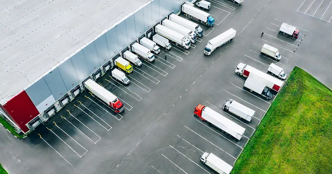 Aerial view of a large distribution center with multiple semi-trucks and trailers parked at loading docks. The warehouse has a gray roof and red accents. The parking area and surrounding green grass are visible.