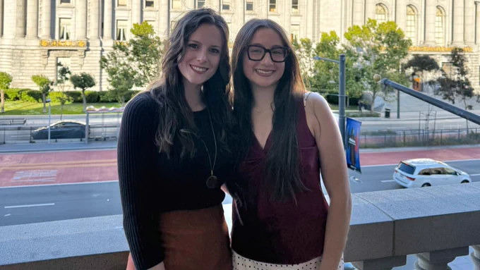 Recent Husker graduates Grace Lewis (left) and Livia Ziskey pose for a photo on a patio overlooking a busy street, with a large government building in the background. (Courtesy)