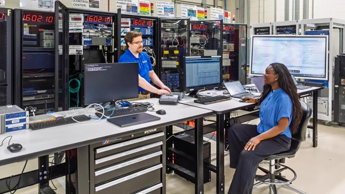 A person is standing and another is sitting in a tech laboratory. They are surrounded by computer monitors, servers, and electronic equipment. 