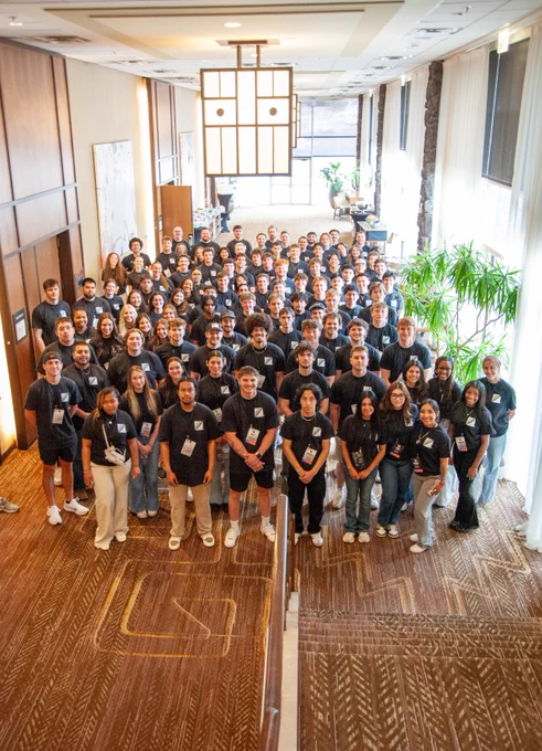 Large group of diverse young adults wearing matching black shirts posing together in a bright, spacious indoor setting with wooden walls and large windows.